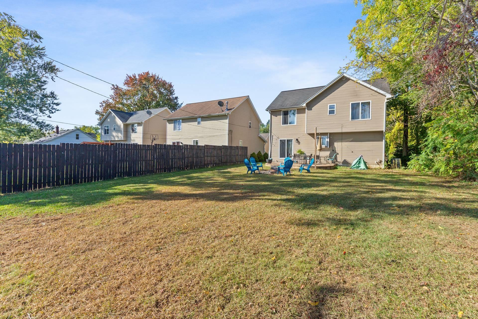 Rear of a tan two-story house with a patio, blue chairs, fenced lawn, and autumn trees.