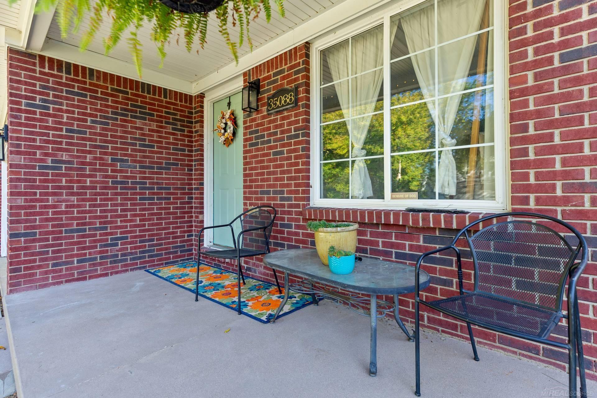 Covered brick front porch with a pale green door, curtained window, and black metal seating.