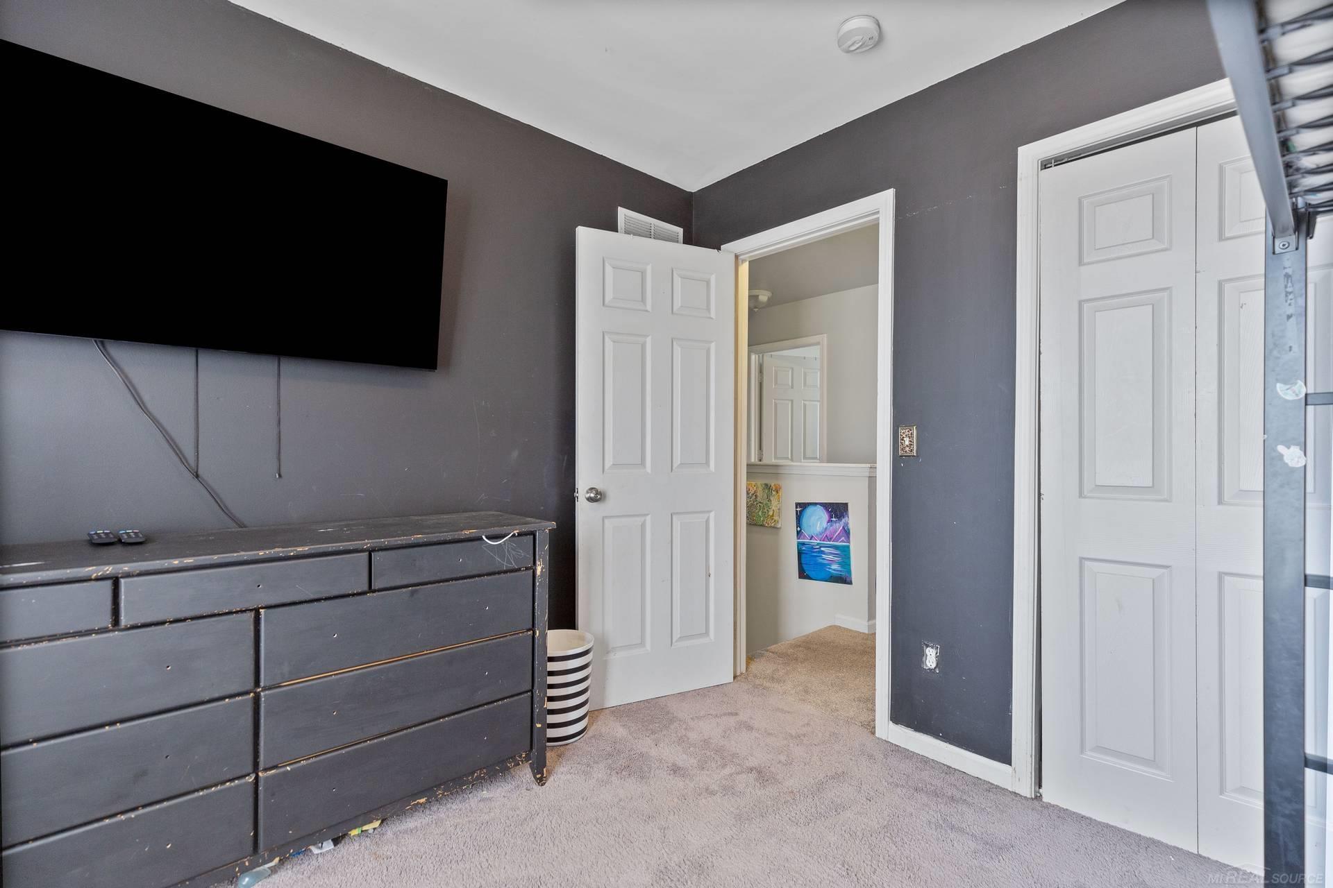 Bedroom with a wall-mounted TV, dark dresser, beige carpet, and white closet doors.