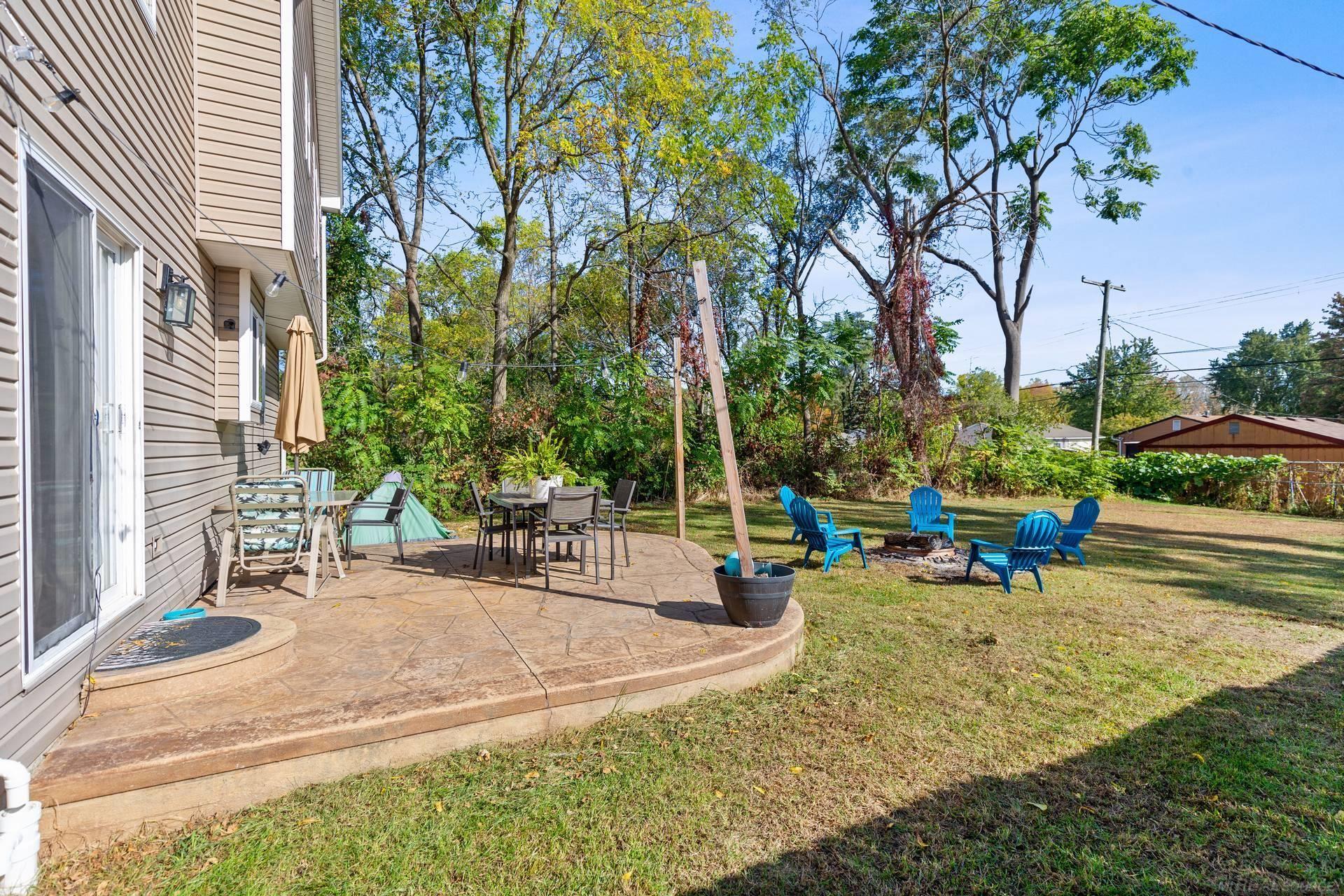 Backyard patio with outdoor dining furniture, blue Adirondack chairs, and a fire pit on the lawn.