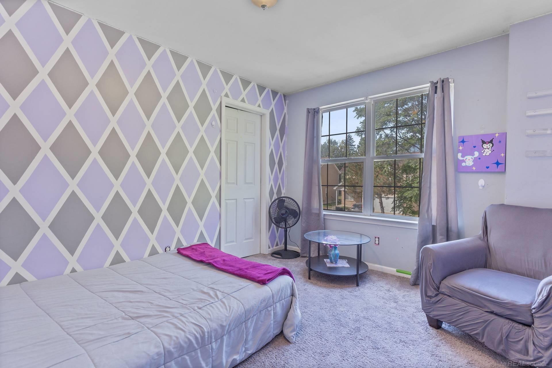 Bedroom with a lavender geometric accent wall, bed, and large curtained window.