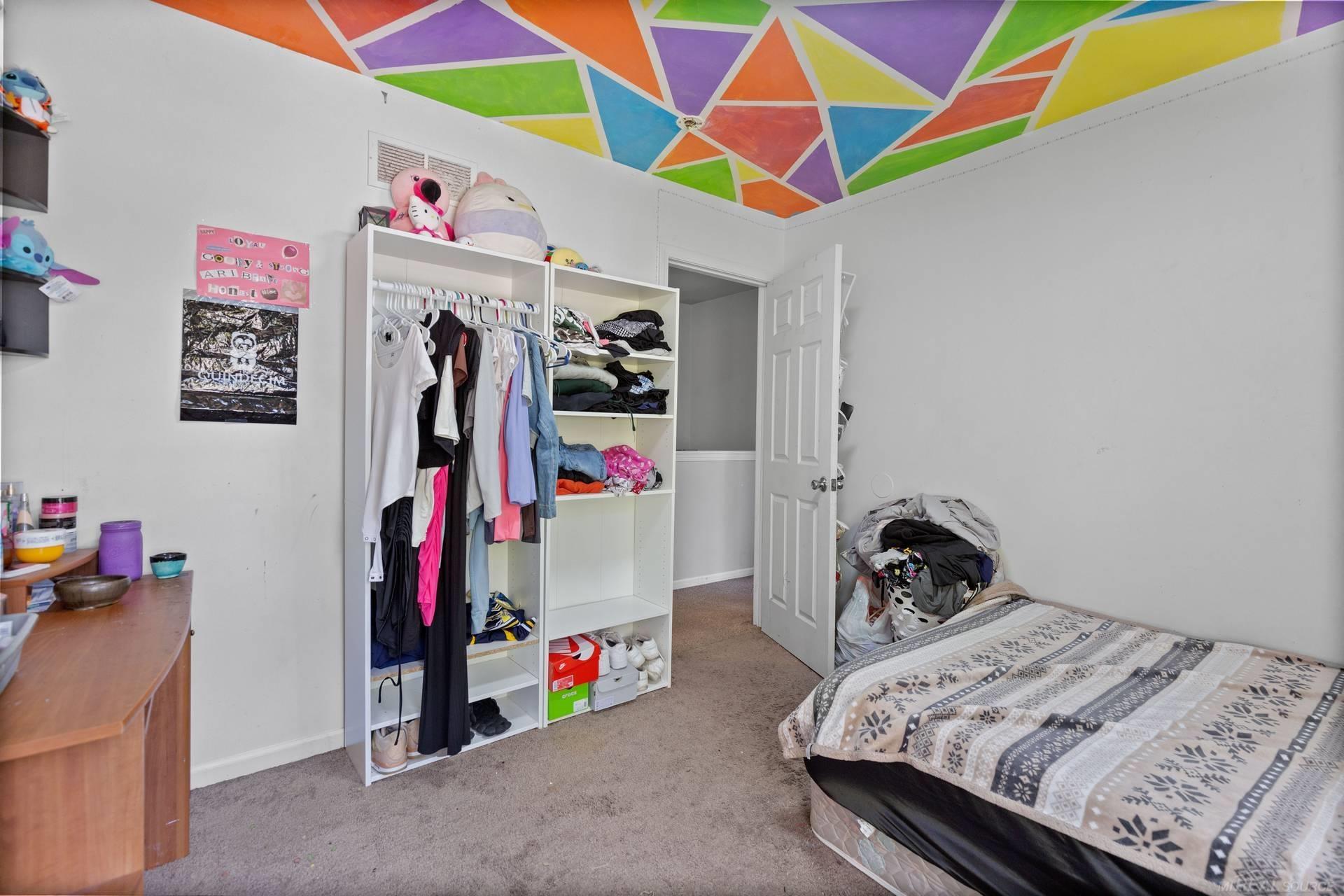 Bedroom with open clothing shelves, a patterned bedspread, and a colorful geometric ceiling.