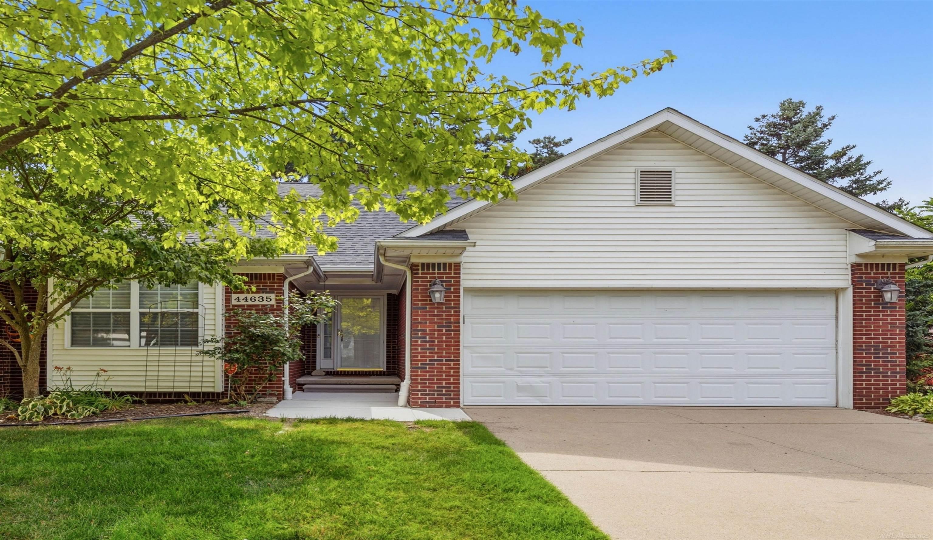 House with white siding, red brick accents, a two-car garage, and a leafy tree over the front lawn.