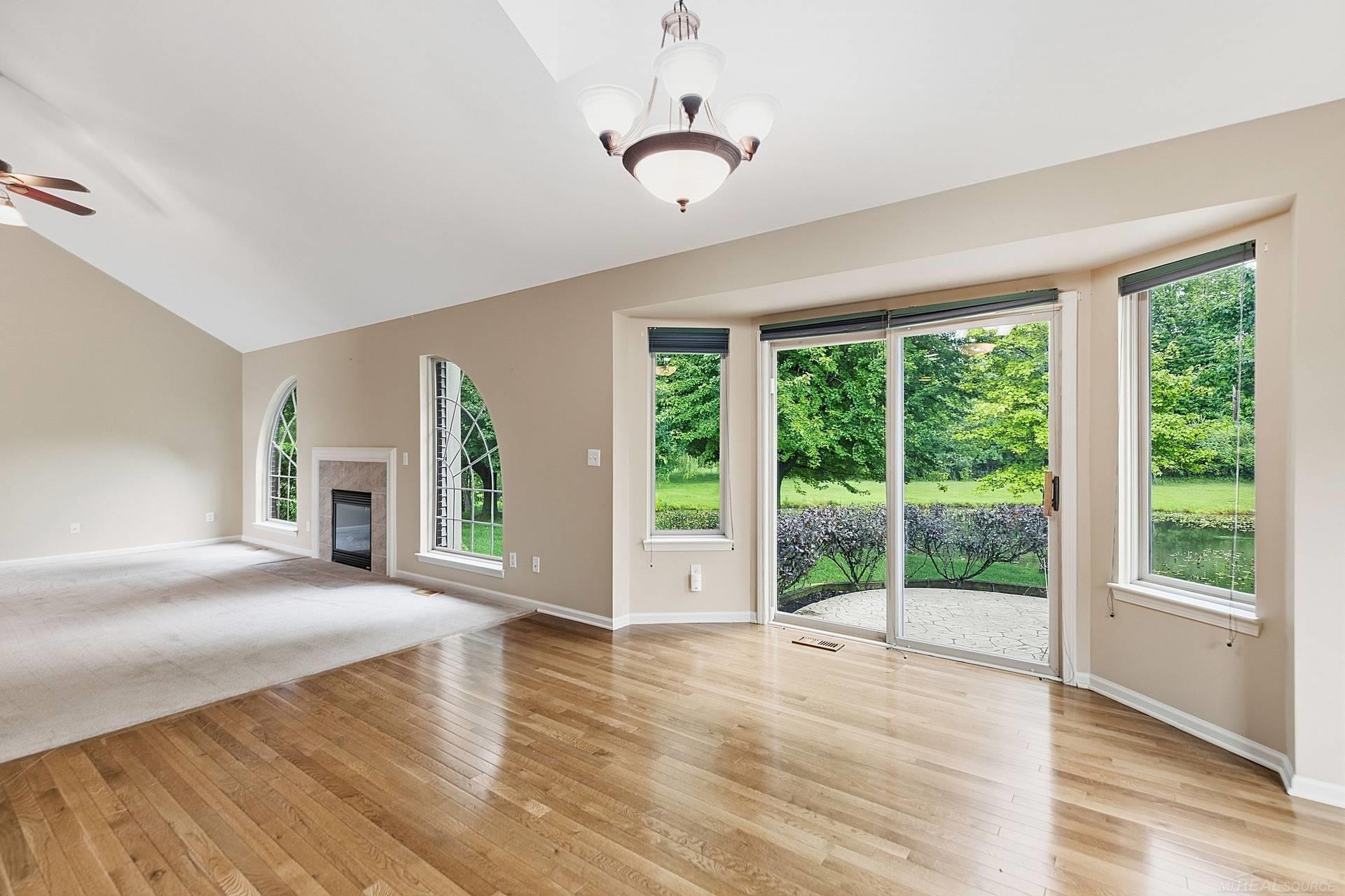 Empty living room with hardwood floors, fireplace, arched windows, and sliding doors overlooking a pond.