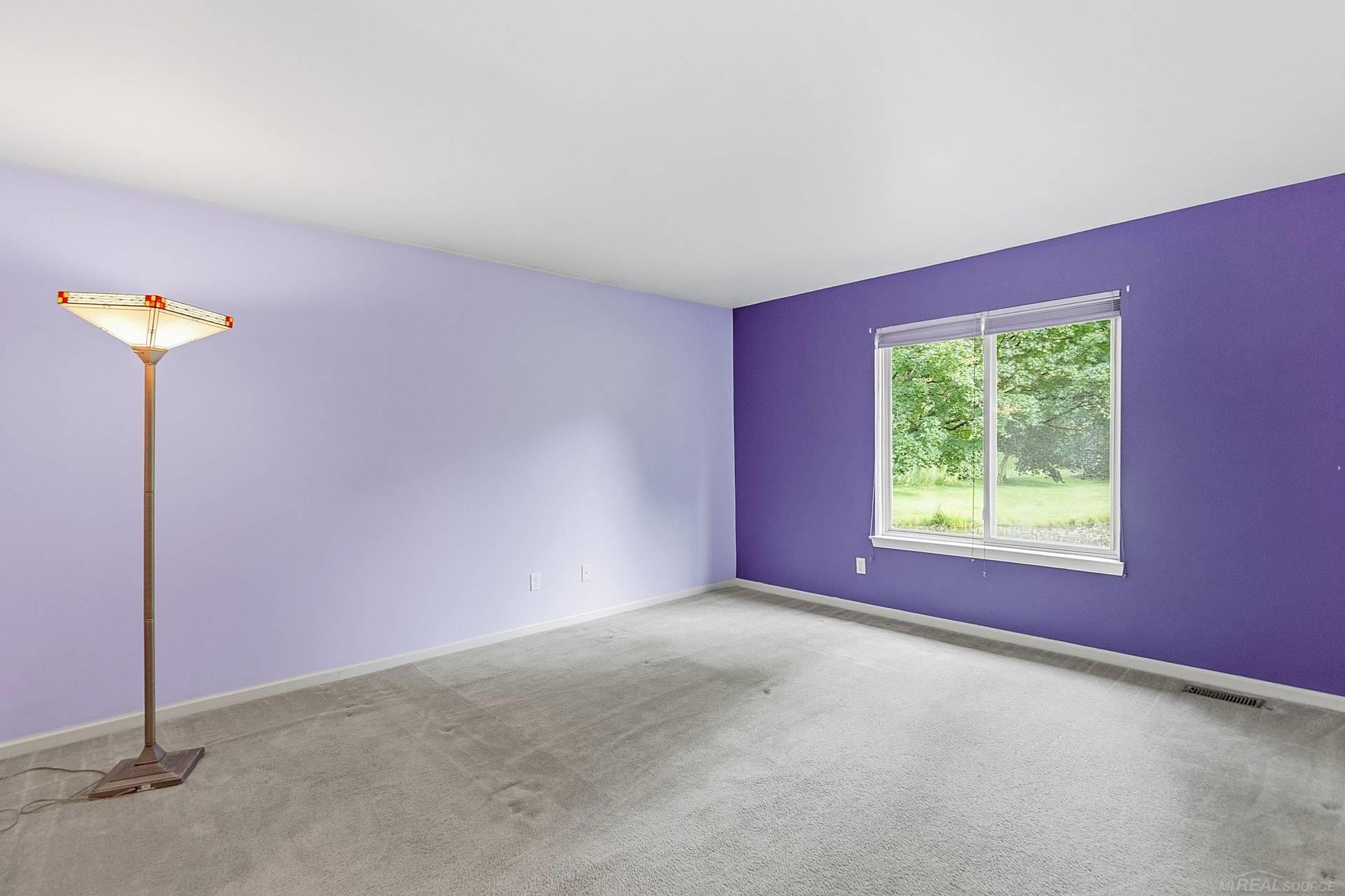 Empty carpeted room with contrasting purple walls, a large window, and a floor lamp.