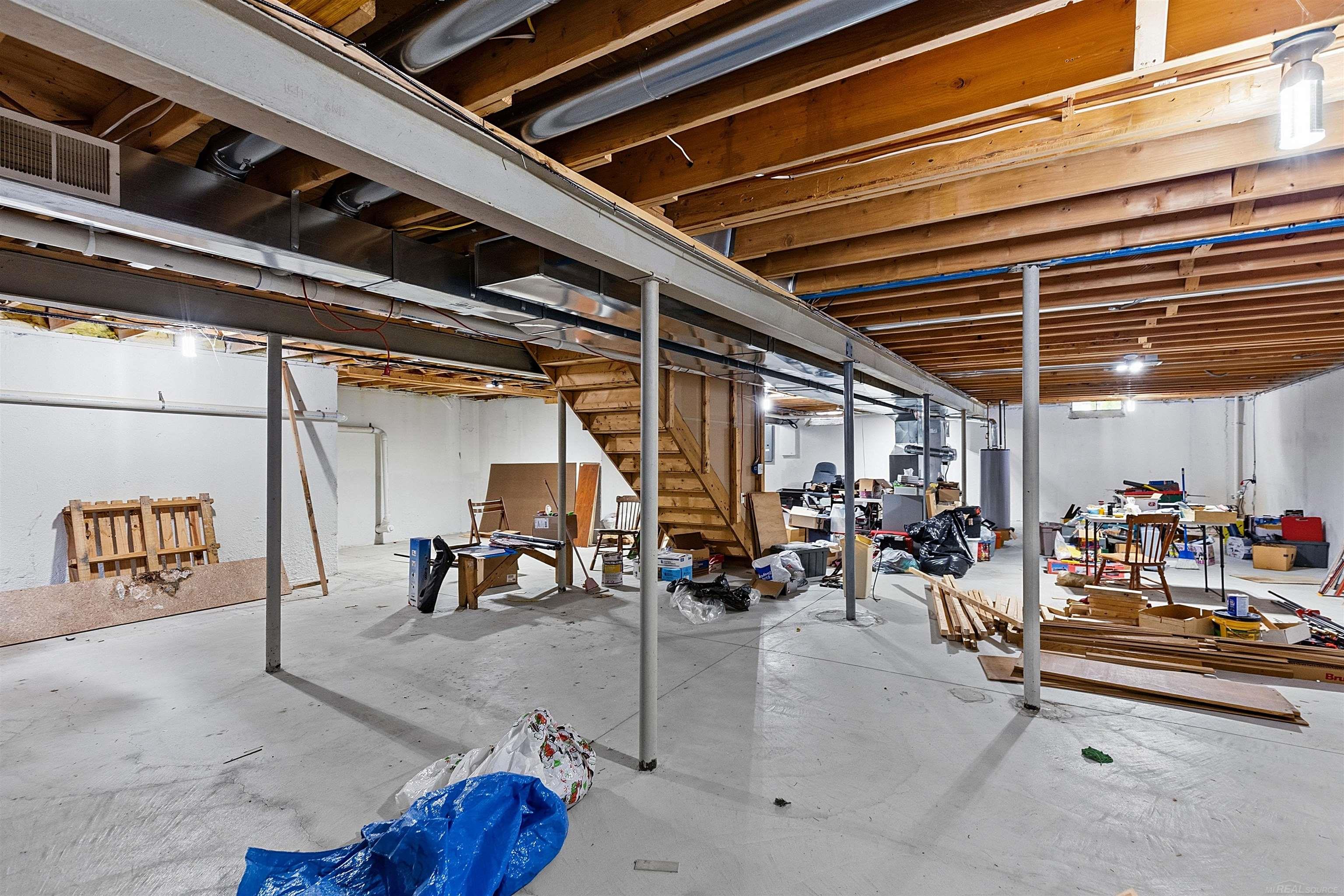 Unfinished basement with exposed joists, support posts, and a staircase amid workshop clutter.