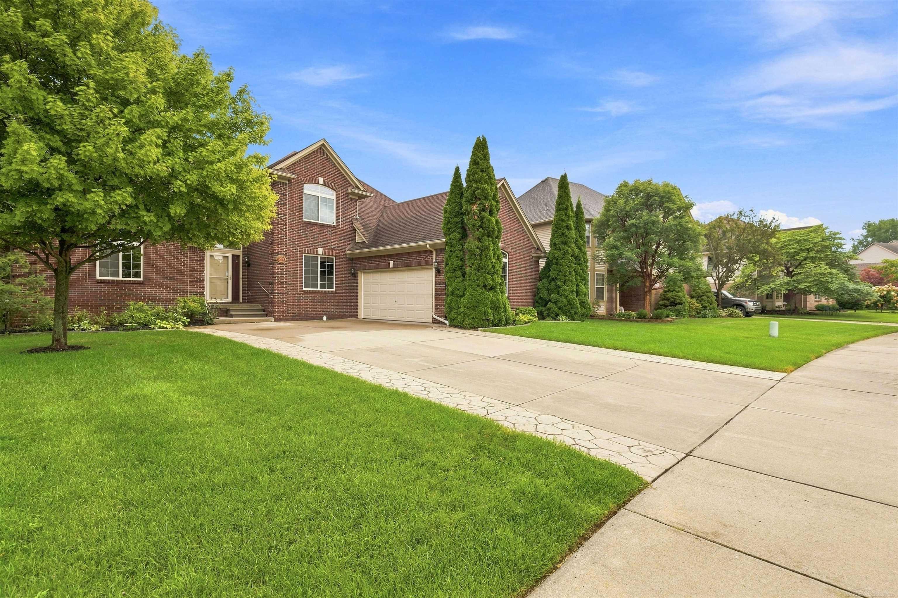 Two-story red-brick house with a tan garage door, wide driveway, and mature trees.