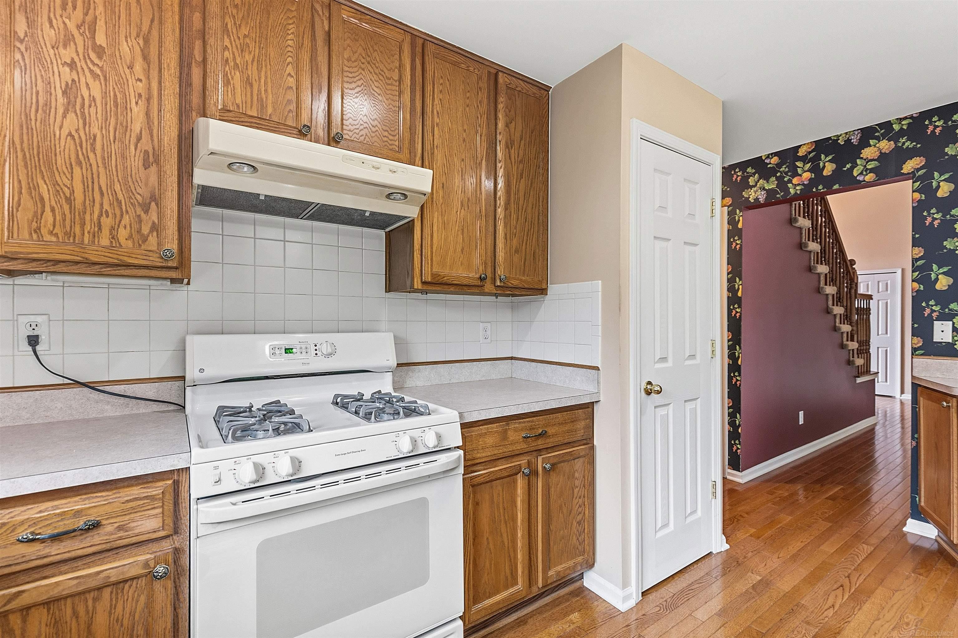 Kitchen with oak cabinets, white tile backsplash, and a white gas range; floral hallway and staircase beyond.
