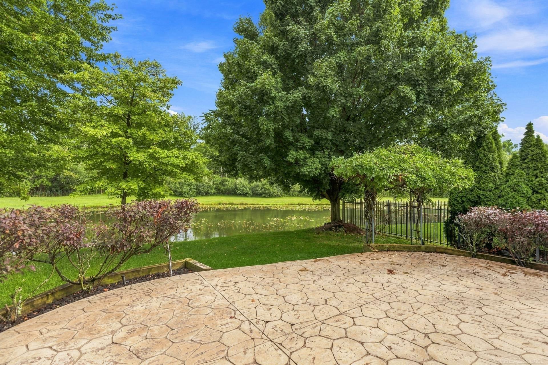 Backyard patio beside a pond, shaded by mature trees and bordered by a metal garden enclosure.