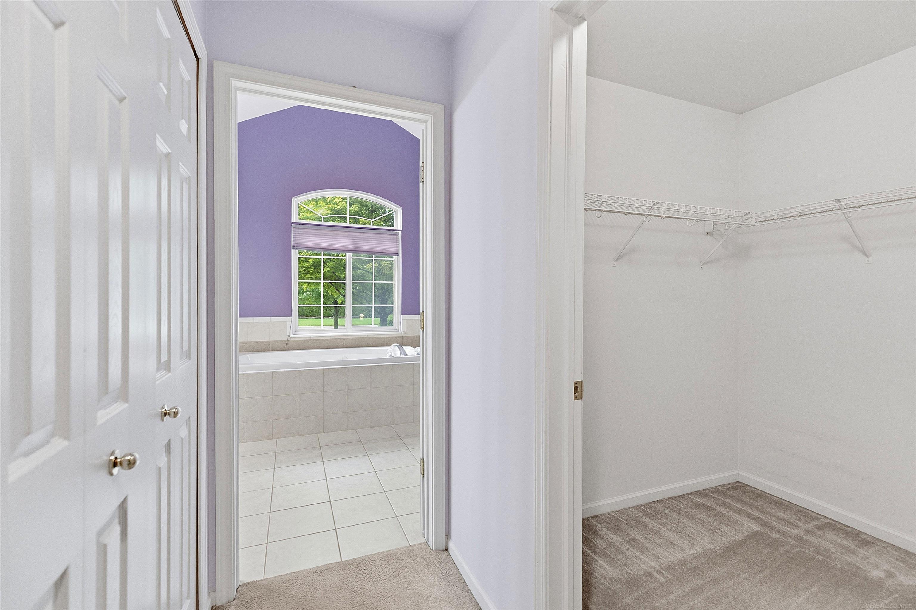 Walk-in closet beside a tiled bathroom with a soaking tub and arched window.