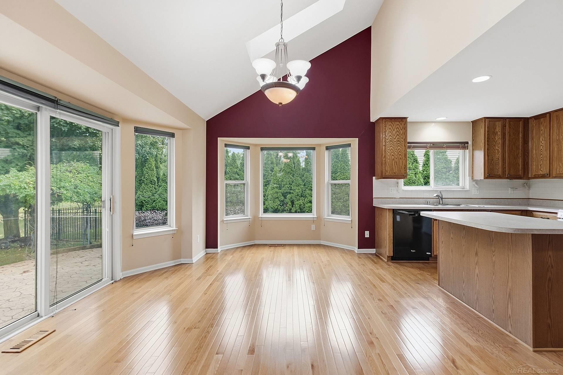 Empty dining area with vaulted ceiling, burgundy accent wall, hardwood floors, and adjoining wood-cabinet kitchen.