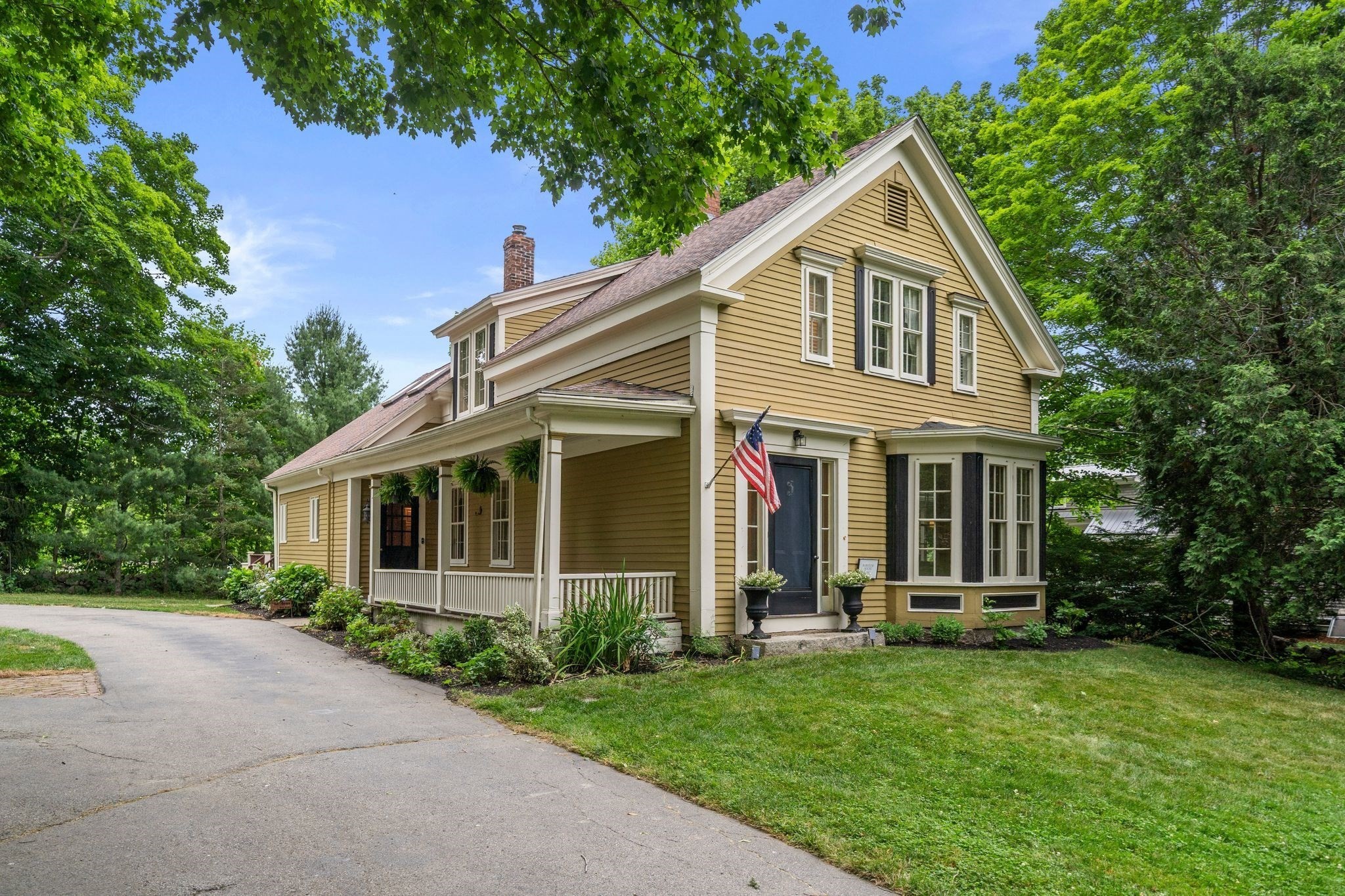 Yellow two-story clapboard house with a covered porch, American flag, leafy trees, and curved driveway.