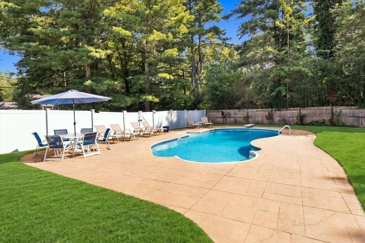 Curved backyard swimming pool bordered by a paved patio, lawn, and patio furniture.