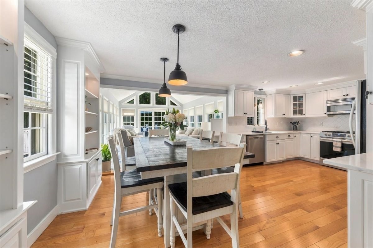 Open kitchen and dining area with white cabinets, a large table, hardwood floors, and a window-lined sunroom.
