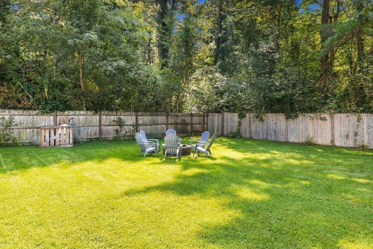 Fenced backyard with a broad green lawn, Adirondack chairs around a fire pit, and mature trees.