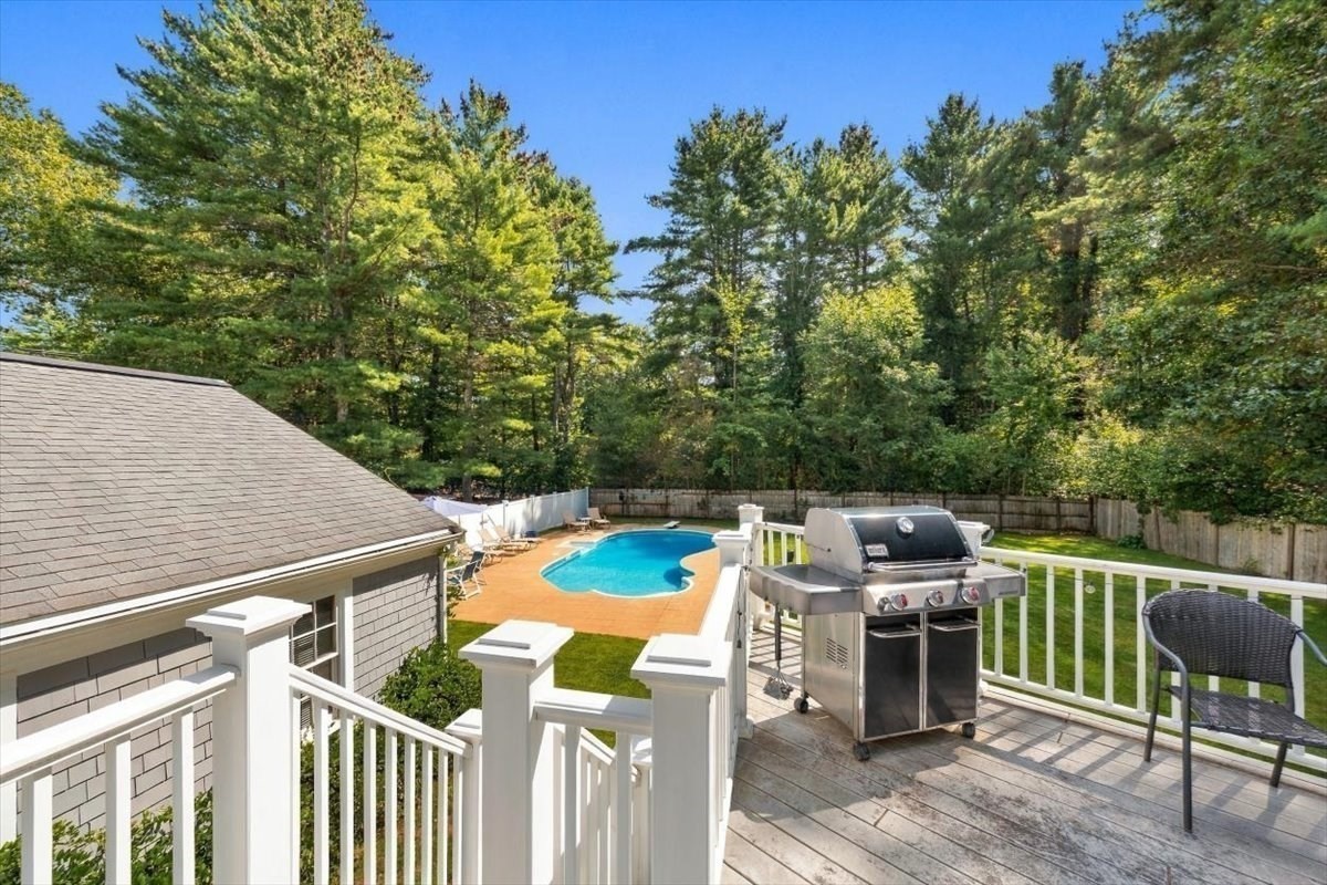 Backyard deck overlooks an in-ground pool, with a stainless-steel grill and mature trees beyond the fence.