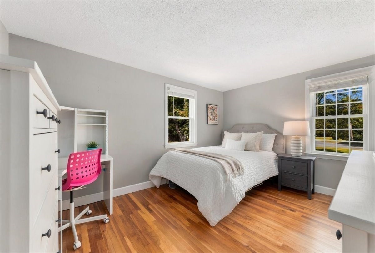 Staged bedroom with a white bed, warm wood floors, and a built-in desk with a pink chair.