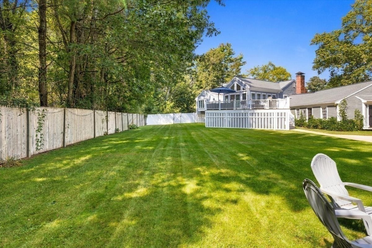 House with a raised deck overlooking a broad fenced lawn shaded by mature trees.