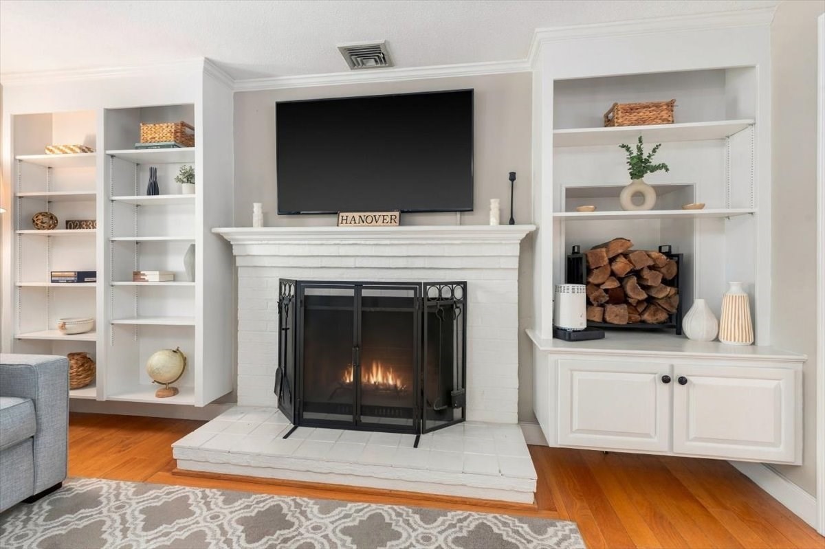 Living room with a white brick fireplace, built-in shelves, mounted TV, and hardwood floors.