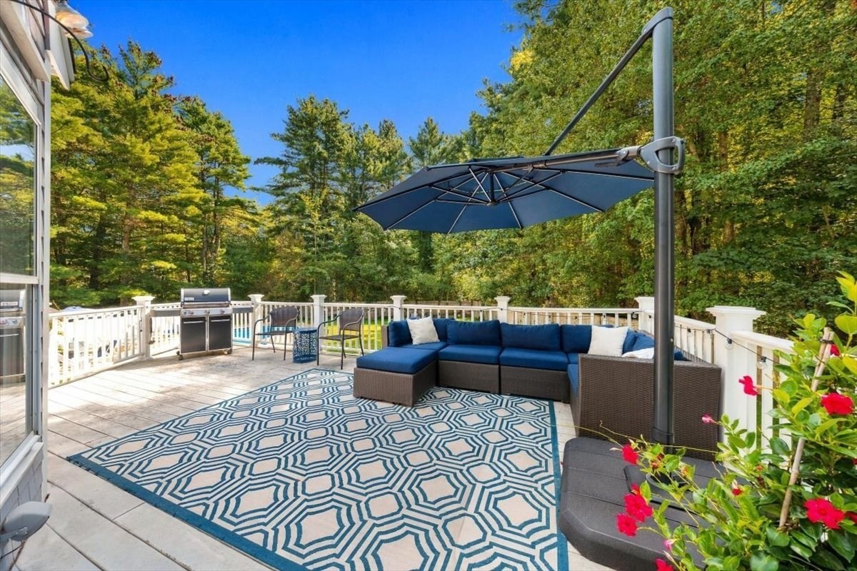 Furnished deck with blue sectional, cantilever umbrella, geometric rug, and grill, surrounded by trees.