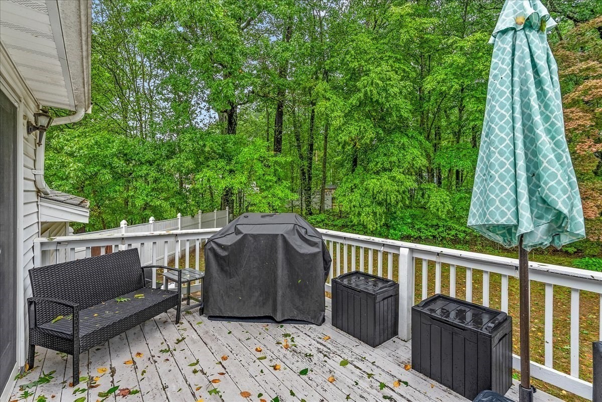 Wooden deck with white railings, a covered grill, wicker bench, and leafy wooded surroundings.