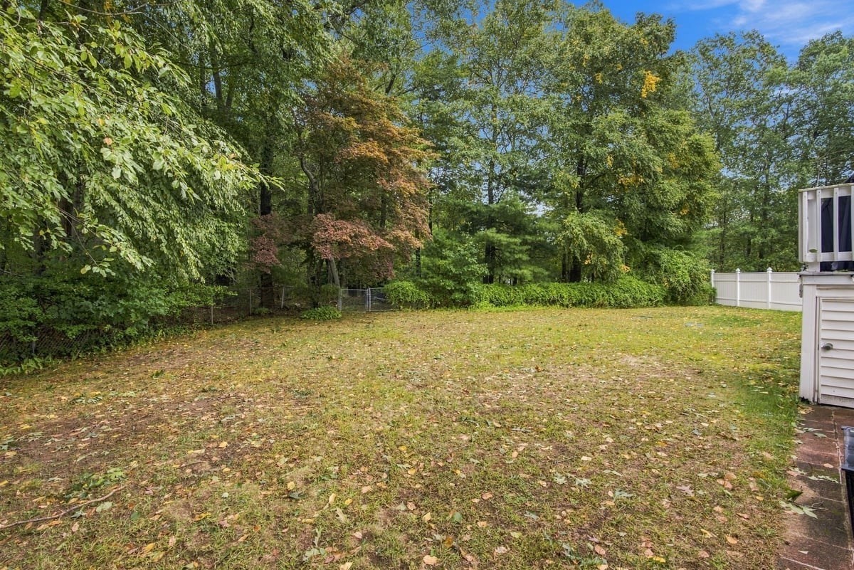 Backyard lawn bordered by mature trees, with a white fence beside the house.