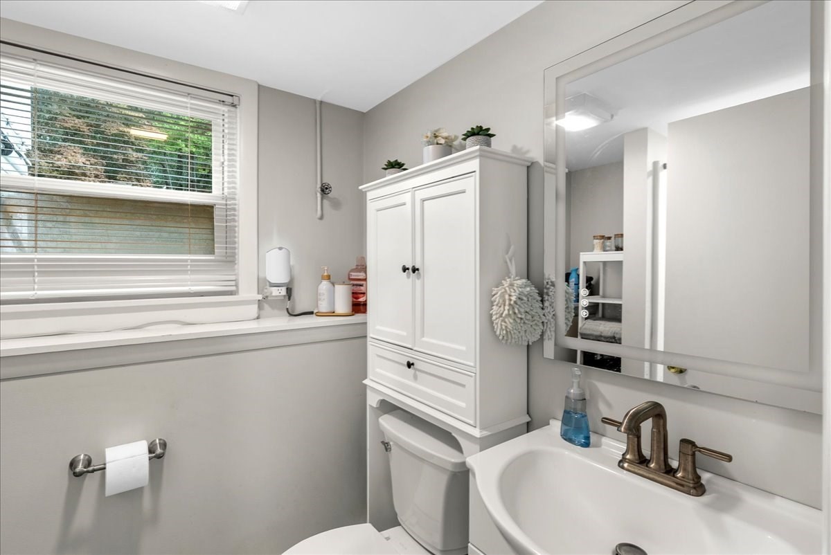 Bathroom with a window-side bathtub, white toilet, tall storage cabinet, and mirrored vanity.