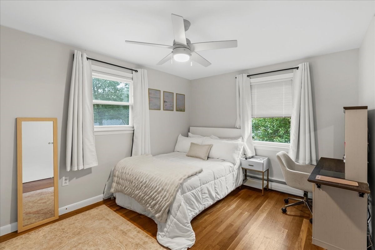 Bedroom with a white bed, two curtained windows, hardwood floors, and a ceiling fan.
