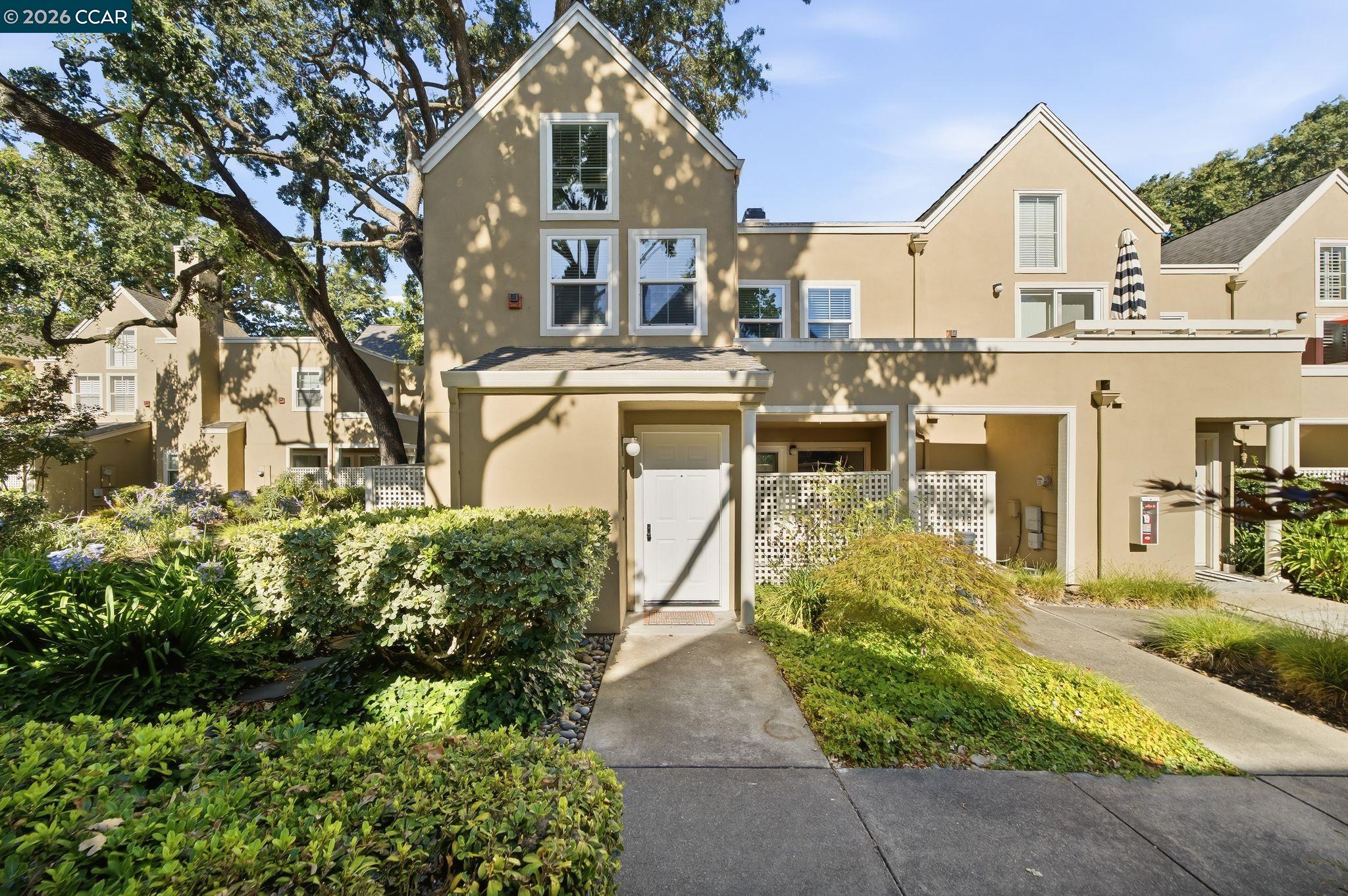 Tan stucco residence with gabled rooflines, landscaped shrubs, and a concrete walkway to the entrance.