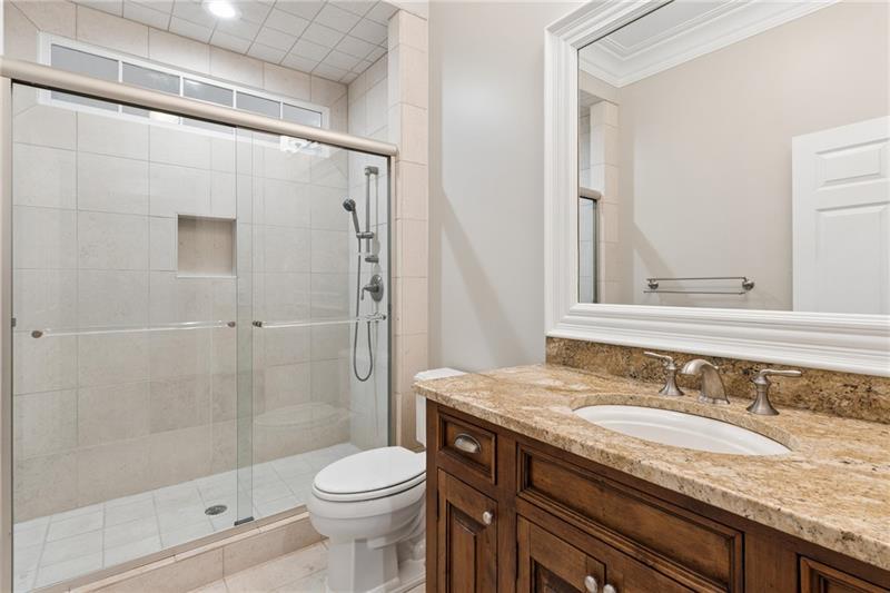 Bathroom with a glass-enclosed tiled shower and dark wood vanity topped with tan stone.