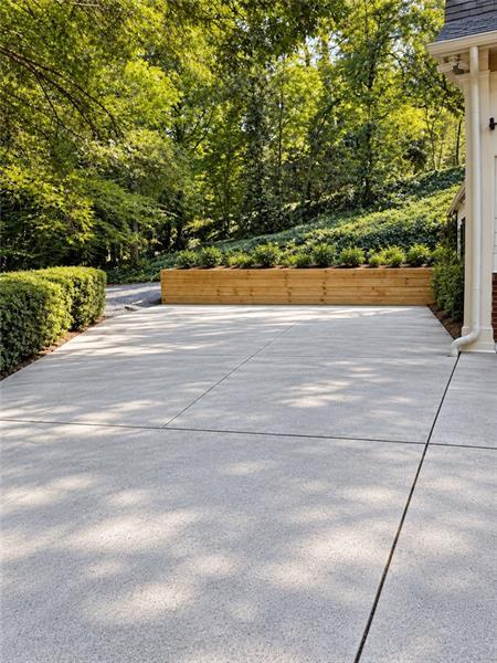 Paved patio beside a house, with a low wooden planter and leafy trees.