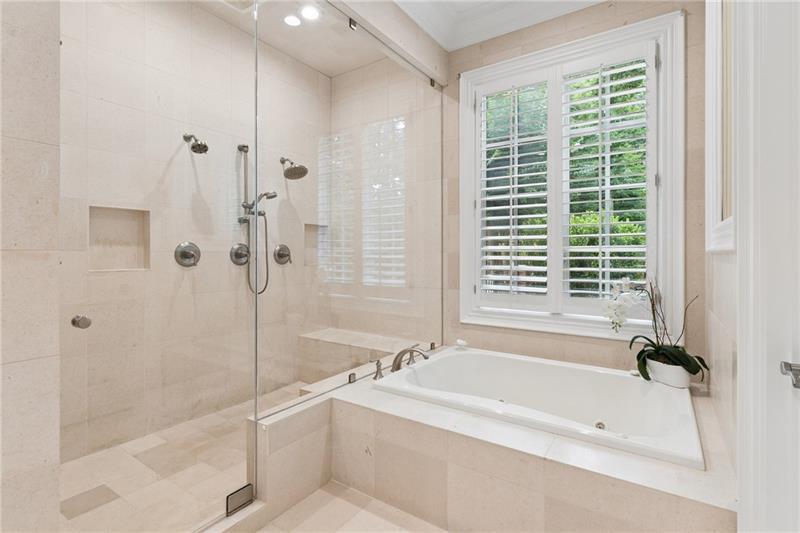 Bathroom with a glass-enclosed tiled shower and corner soaking tub beneath shuttered windows.