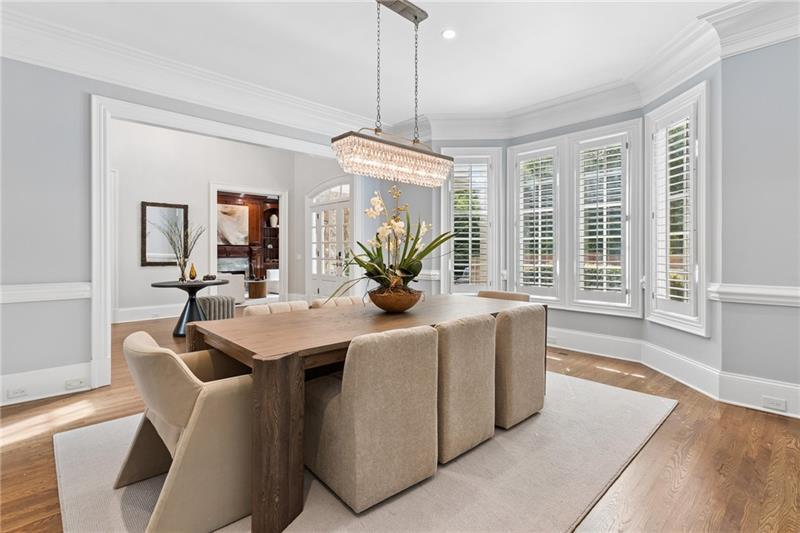 Dining room with a wooden table, upholstered chairs, crystal chandelier, and shuttered bay windows.
