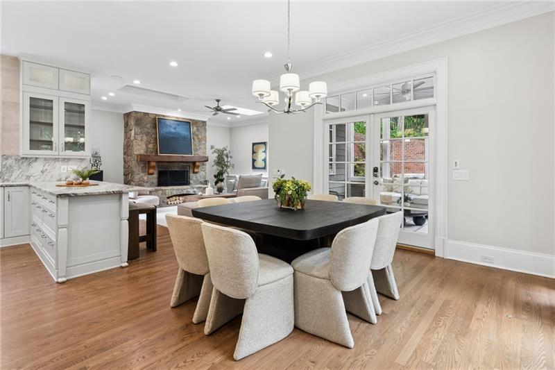 Open-plan dining room with a dark table, upholstered chairs, white kitchen cabinetry, and an adjoining stone fireplace.