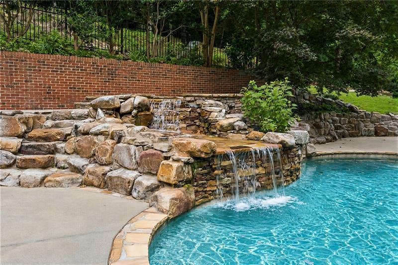 Backyard pool with cascading stone waterfalls, a brick retaining wall, and mature trees.