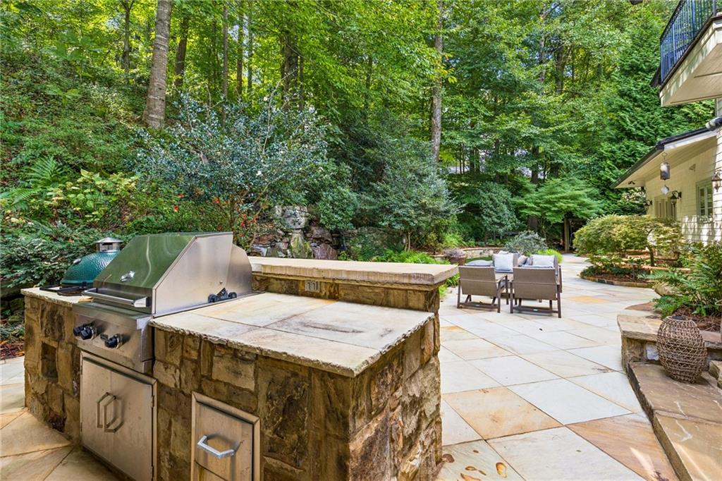 Stone outdoor kitchen and patio with lounge seating, surrounded by mature trees.