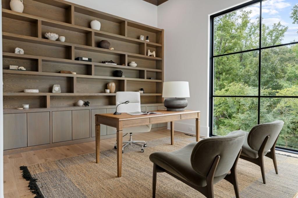 Home office with built-in wood shelving, a desk, upholstered chairs, and a large window overlooking trees.
