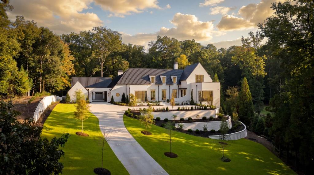 Modern white home with dark metal roofs, landscaped lawn, curved retaining wall, and wooded surroundings.