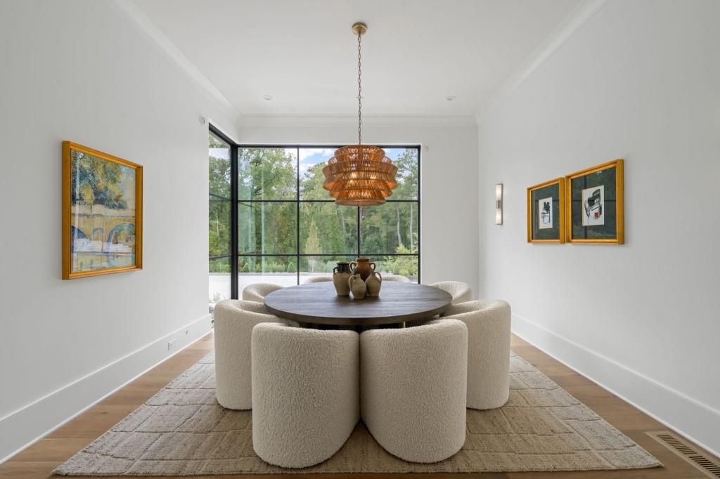 Dining room with a round table, cream chairs, woven pendant, and large windows overlooking trees.
