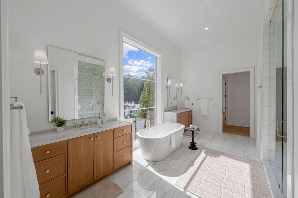 Bright bathroom with a freestanding tub, tall window, marble floors, and wood vanities.