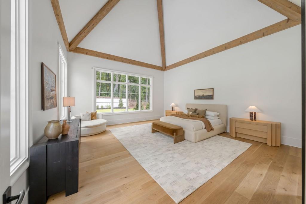 Staged bedroom with a vaulted timber-beam ceiling, large windows, and a bed on a pale rug.