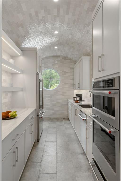 Bright narrow kitchen with an arched tiled ceiling, round window, and stacked stainless steel ovens.