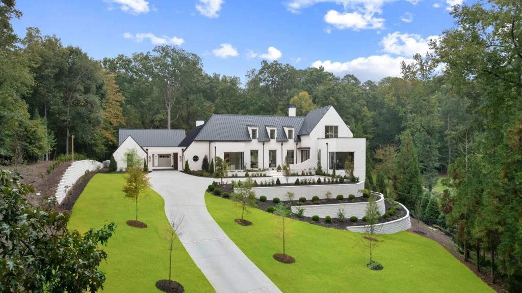 Modern white home with a dark metal roof, landscaped terraces, and a broad driveway in a wooded setting.