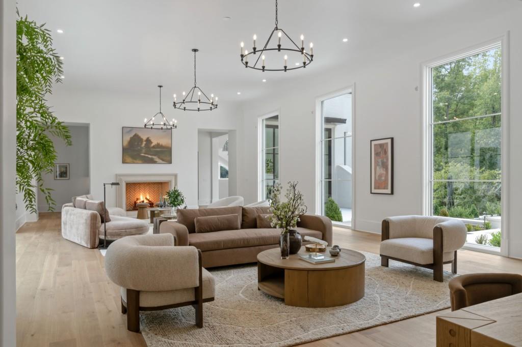 Staged living room with a fireplace, neutral seating, three chandeliers, and tall windows overlooking trees.