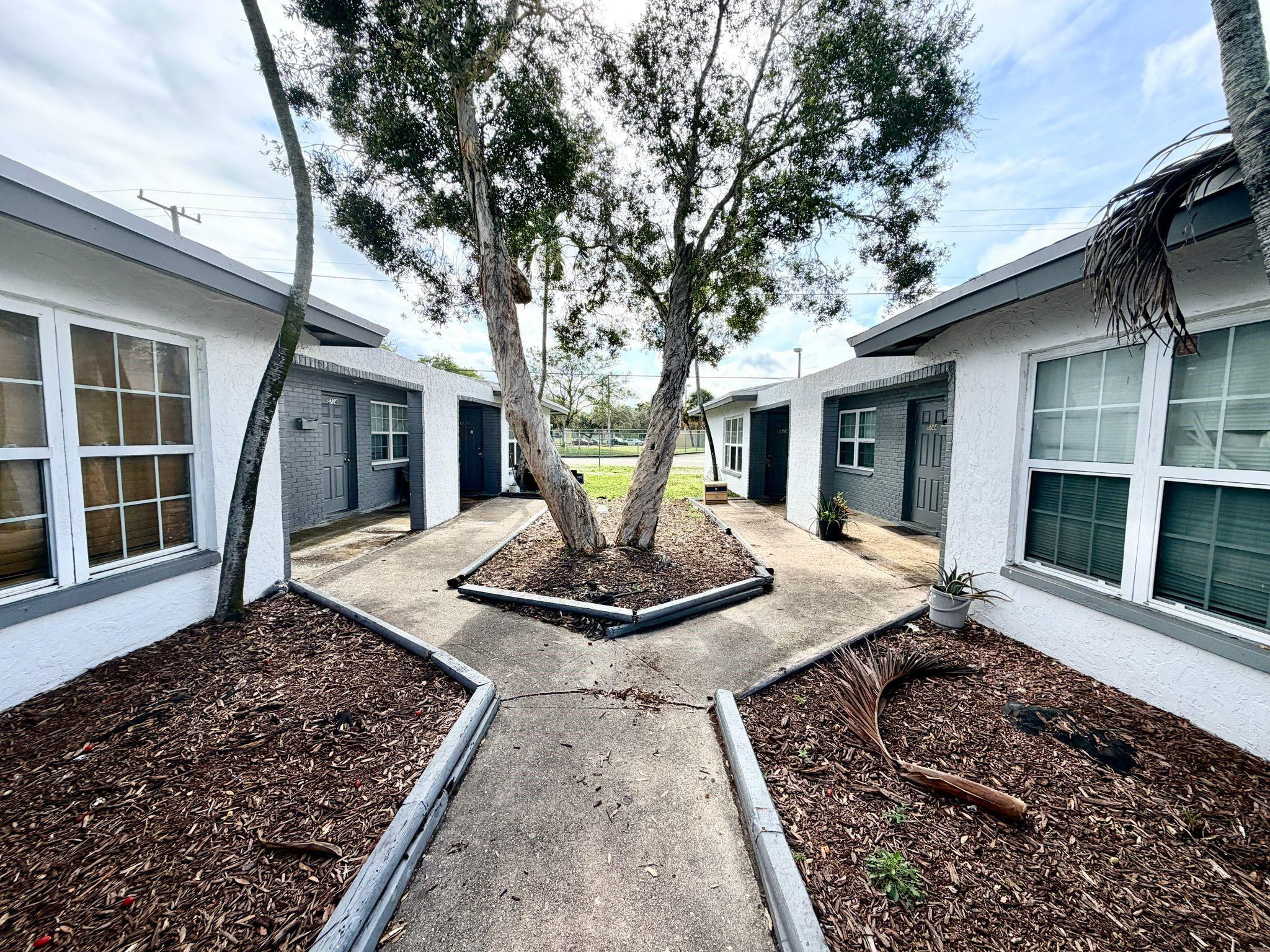 Residential courtyard with a paved path winding around mature trees between two low-rise buildings.