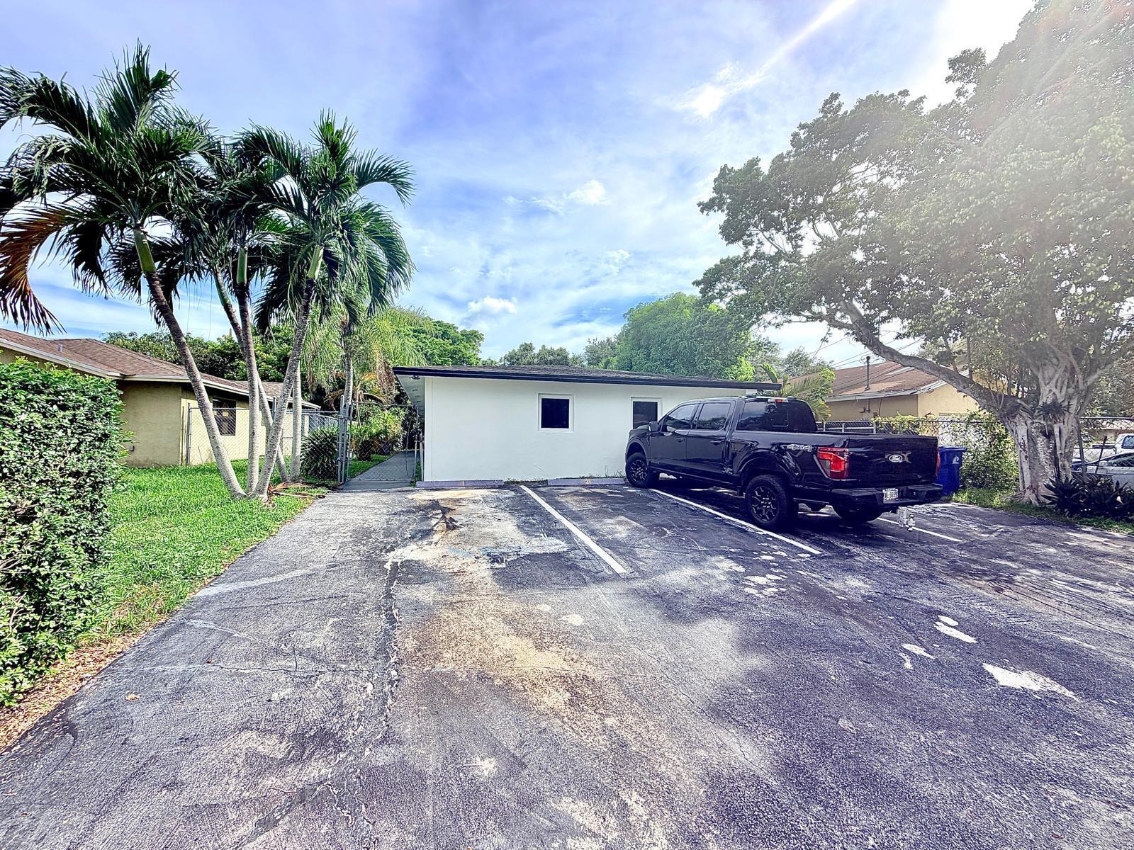 White single-story building beside a paved parking area, with a black pickup truck and palm trees.