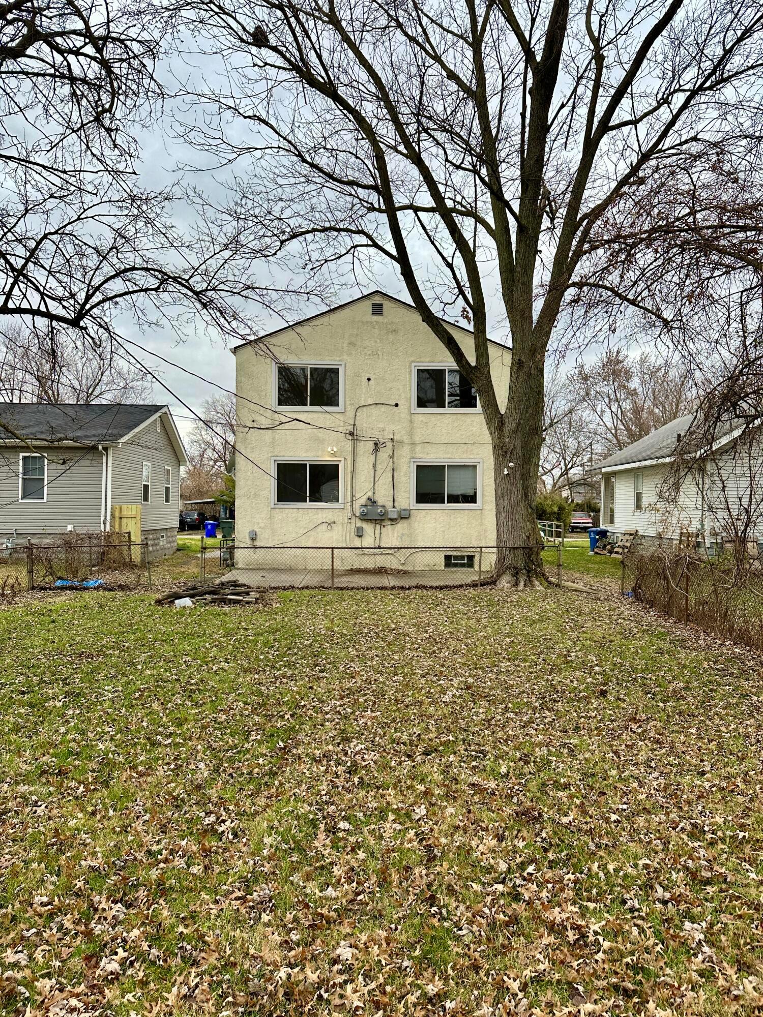 Rear of a two-story cream house behind a leaf-covered yard, with a large tree and chain-link fence.