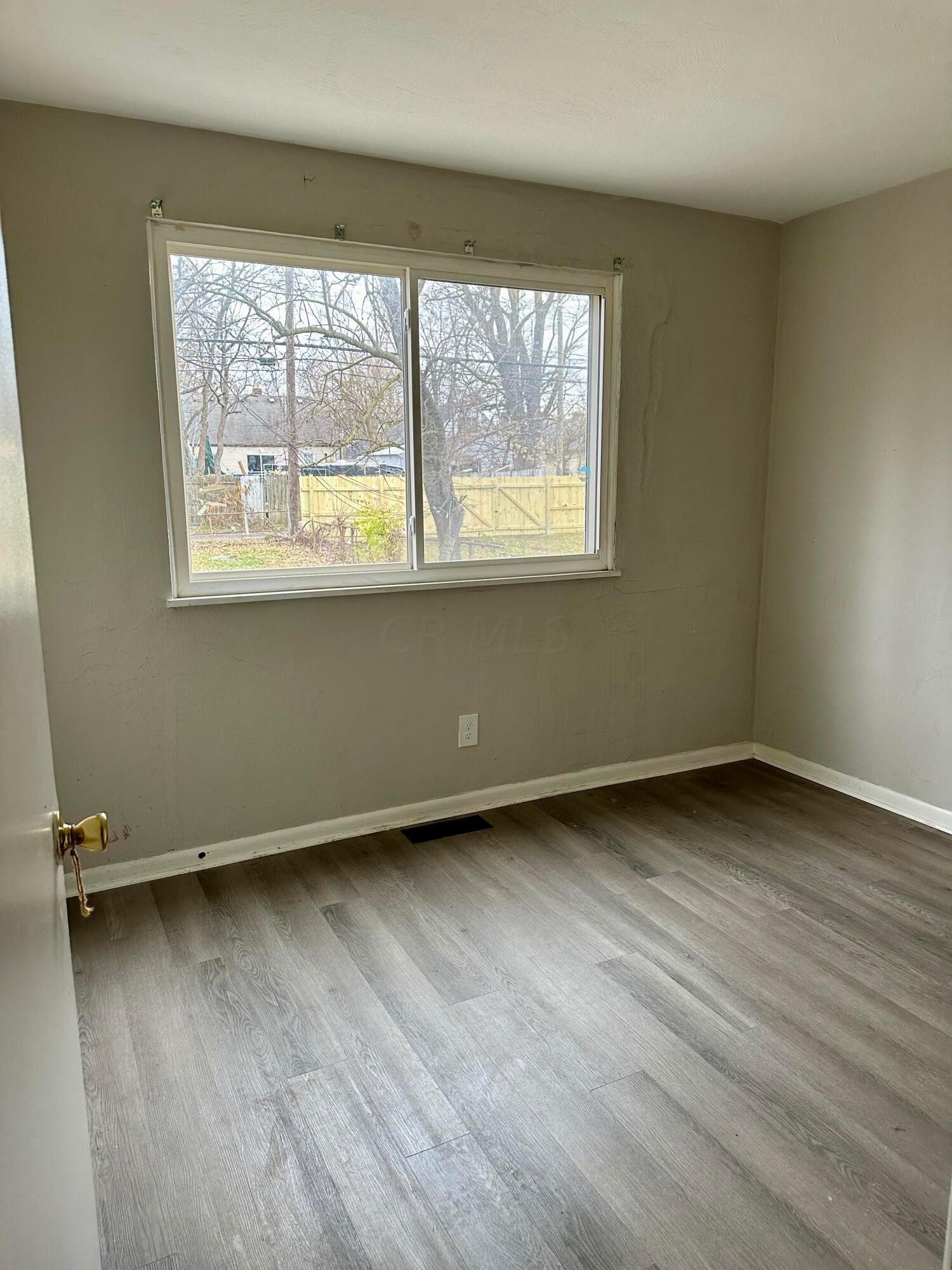 Vacant room with gray wood-look flooring and a large window overlooking a fenced yard.