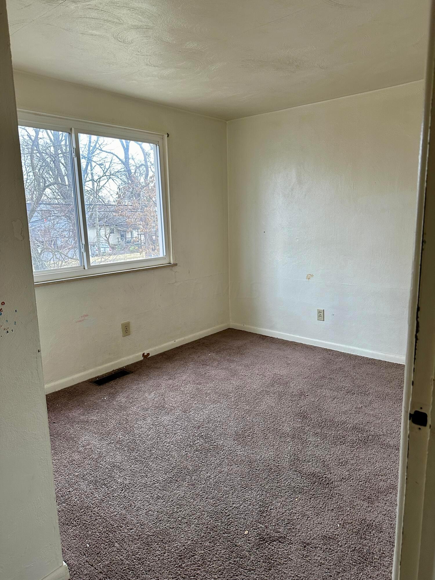 Empty bedroom with worn brown carpet and a large window overlooking leafless trees.