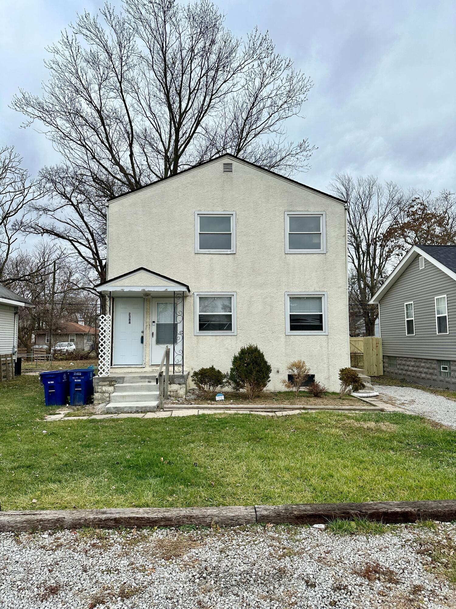 Two-story white stucco duplex with a small covered porch and gravel driveway.